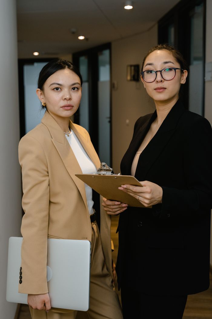 Two confident businesswomen in suits holding laptop and clipboard in an office setting.