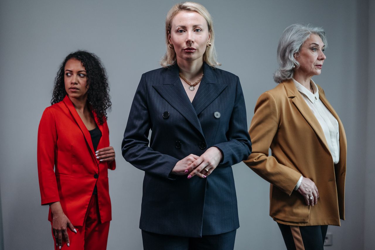 About Three businesswomen in suits pose confidently on a neutral background, showcasing diversity.